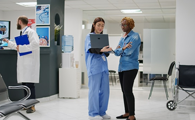 Duas mulheres em pé conversando na recepção de um hospital. A da esquerda é uma doutora com um laptop, mostrando para a paciente na direita.